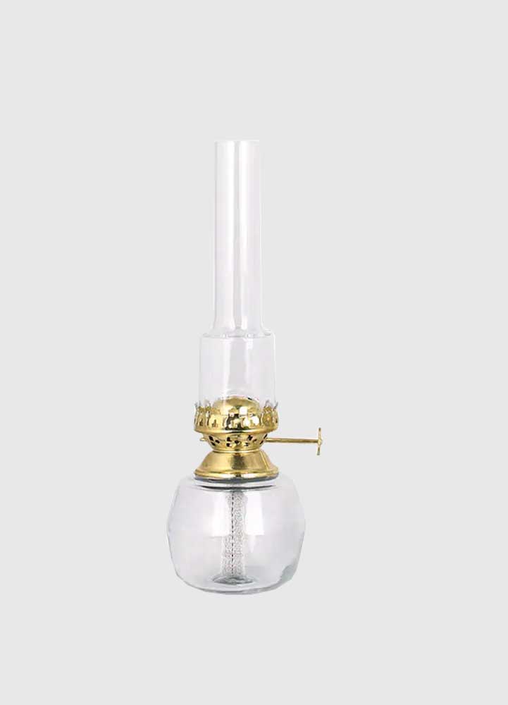 Hög oljelampa i klart ofärgat klarglas med mässingdetaljer och brännarrör i klart färgat glas placerad på en vit bakgrund.
