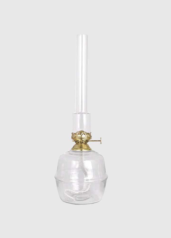 Hög oljelampa i klart ofärgat klarglas med mässingsdetaljer och högt brännarrör i klart färgat glas placerad på en vit bakgrund.
