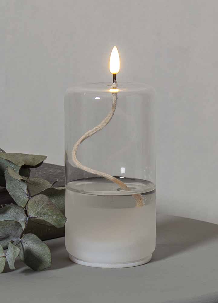 Oljelampa i genomskinligt glas med en konstgjord låga på batteri, oljelampan står på ett grått bord med en eukalyptuskvist bredvid. 
